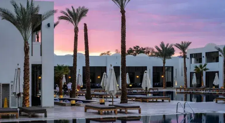 Modern white hotel resort with pool, palm trees, and white umbrellas at sunset in Greece