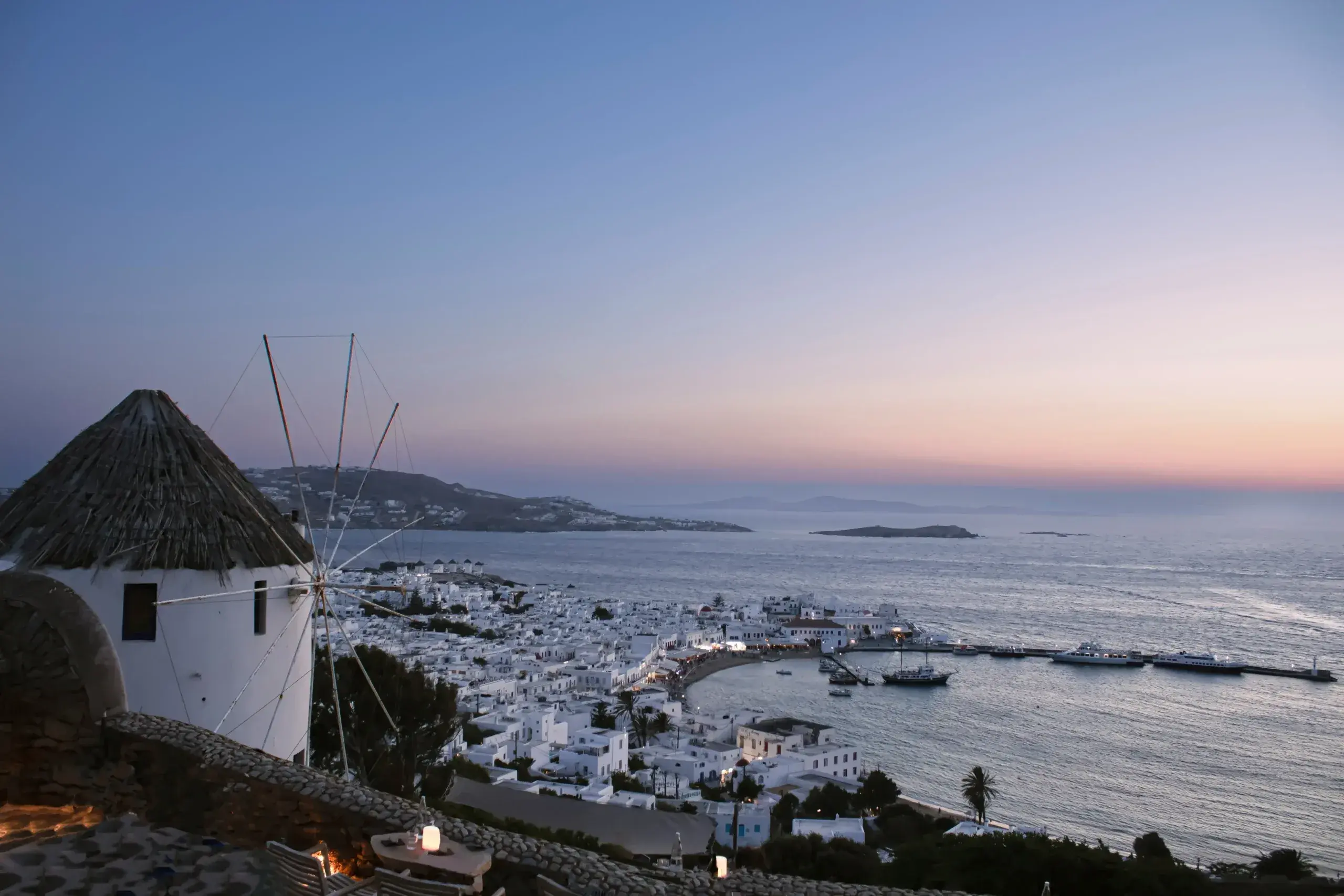 Iconic white windmill with thatched roof overlooking Mykonos town, whitewashed buildings, harbor with boats, and Aegean Sea at dusk